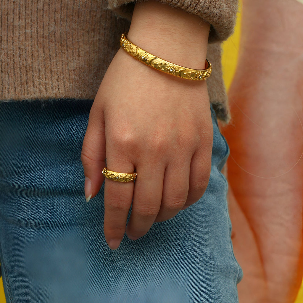 Bracelet jonc fin doré en acier inoxydable gravé de motifs astres porté au poignet, bague assortie au doigt.