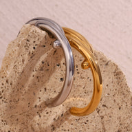 Bracelet jonc entrelacé en acier inoxydable, modèle Jovana, finition argent et or brillant sur pierre beige textured.