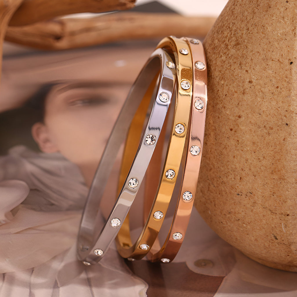 Trois bracelets joncs en acier inoxydable or, or rosé et argent avec éclats de lumière circulaires.