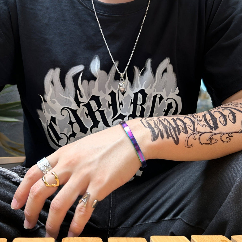 Bracelet jonc fin en acier inoxydable arc-en-ciel brillant porté sur poignet masculin avec tatouage et bagues argentées.
