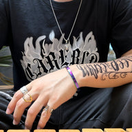 Bracelet jonc fin en acier inoxydable arc-en-ciel brillant porté sur poignet masculin avec tatouage et bagues argentées.