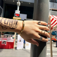 Bracelet jonc en acier inoxydable doré, finition lisse et moderne, porté au poignet masculin tatoué avec plusieurs bagues en .