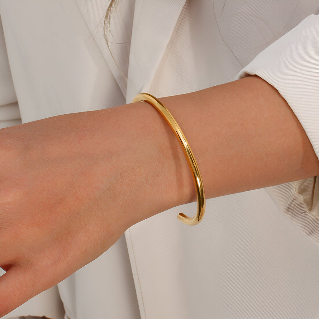 Bracelet jonc en acier inoxydable doré au design épuré porté sur poignet féminin avec veste blanche.