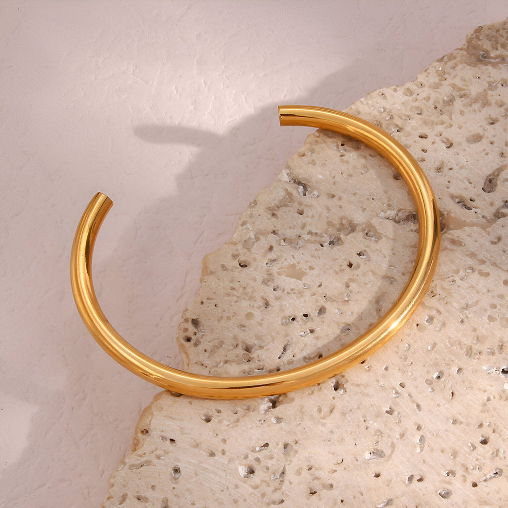 Bracelet jonc en acier inoxydable doré au design épuré posé sur pierre texturée beige.