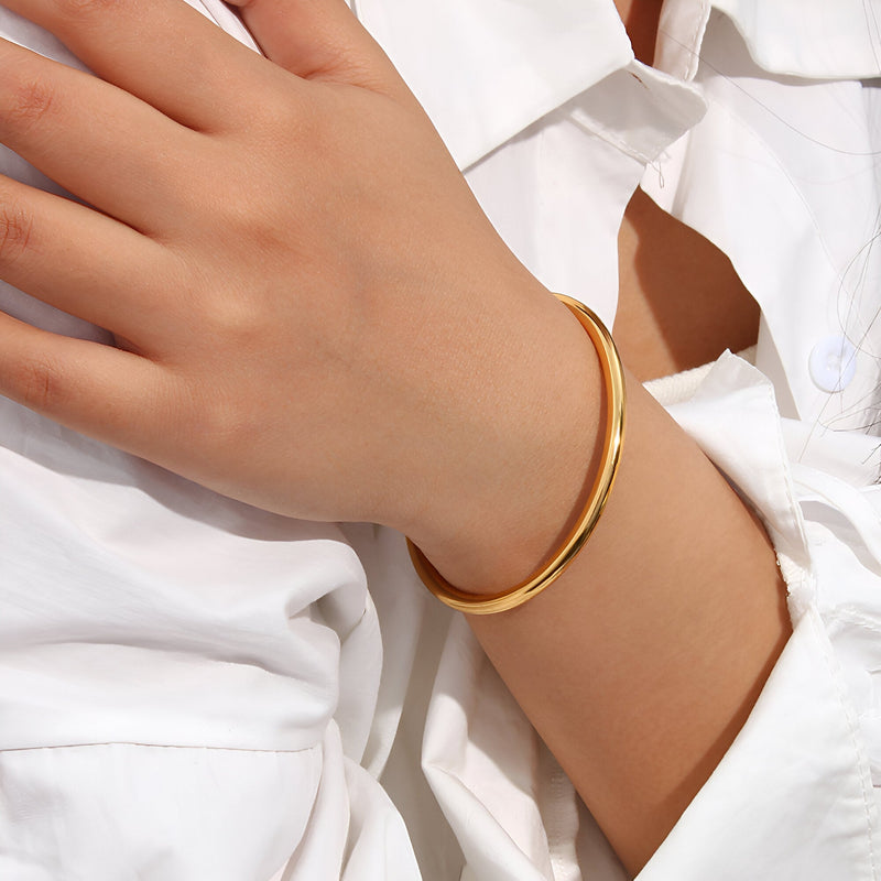 Bracelet jonc fin en acier inoxydable doré, style épuré porté au poignet féminin avec chemise blanche.