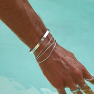 Bracelet jonc argenté en acier inoxydable brillant porté au poignet d’un homme, au-dessus de l’eau turquoise.