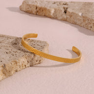 Bracelet jonc doré en acier inoxydable avec finition martelée posée sur pierres beiges texturées.