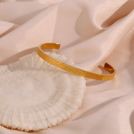 Bracelet jonc or en acier inoxydable à finition martelée élégante posé sur tissu beige doux.