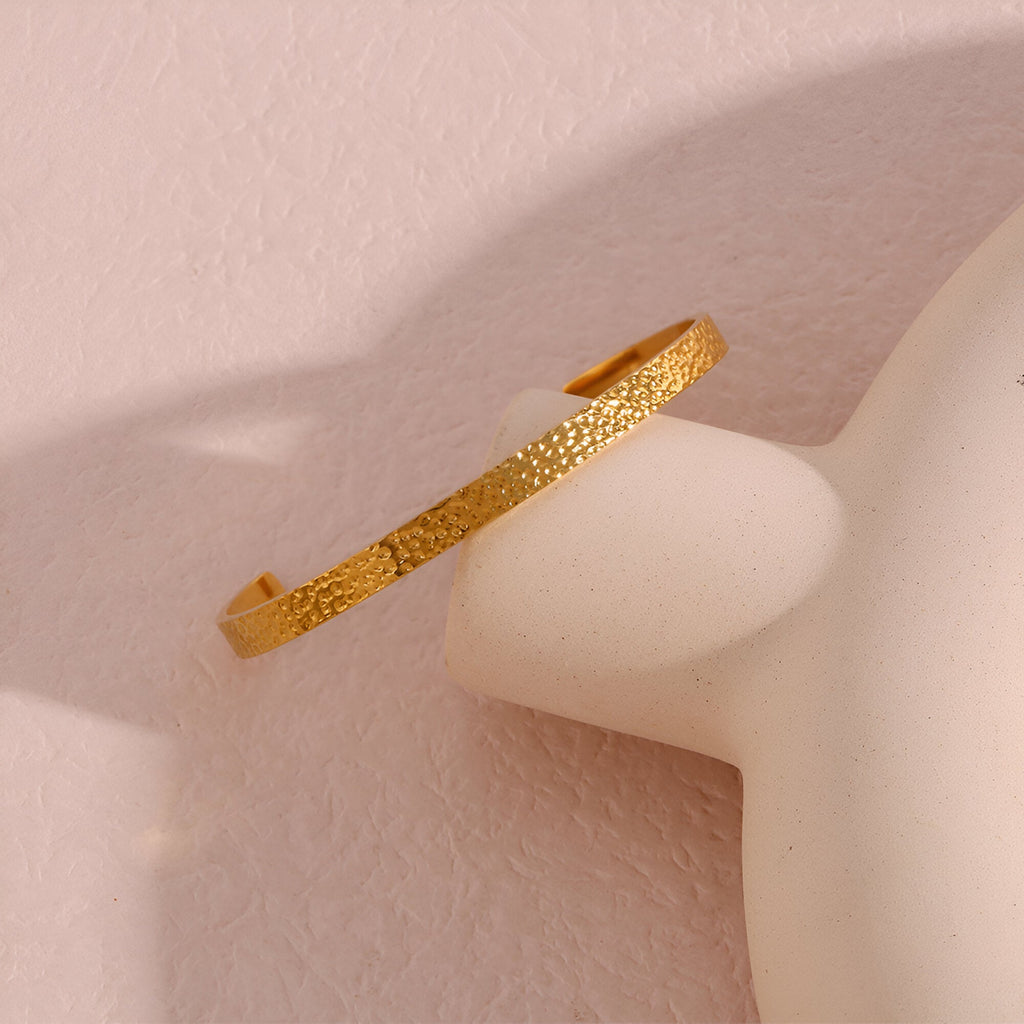 Bracelet jonc fin martelé en acier inoxydable doré posé sur un support blanc lisse.