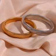 Bracelets jonc flexibles en maille acier inoxydable, un or brillant et un argent mat posés sur tissu rose pâle.