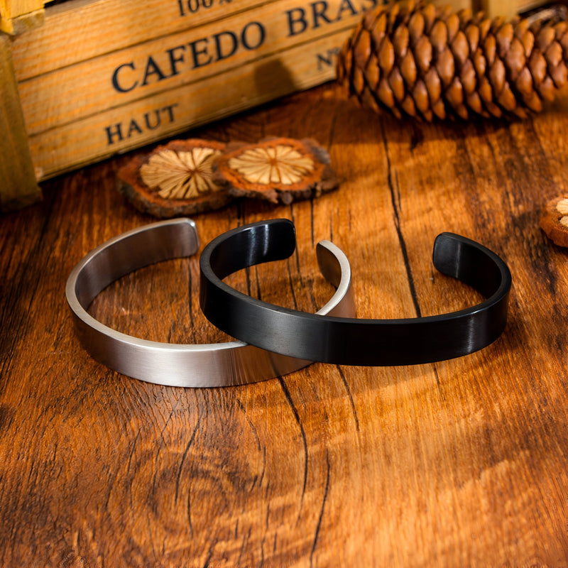 Deux bracelets joncs en acier inoxydable, un argenté brossé et un noir mat, posés sur surface en bois.