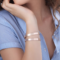 Bracelet jonc en or rosé avec gravures personnalisées sur peau féminine portant une chemise bleue à rayures.