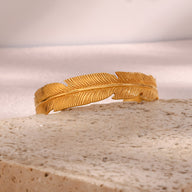 Bracelet jonc en acier inoxydable doré avec motif plume délicate et finition texturée.