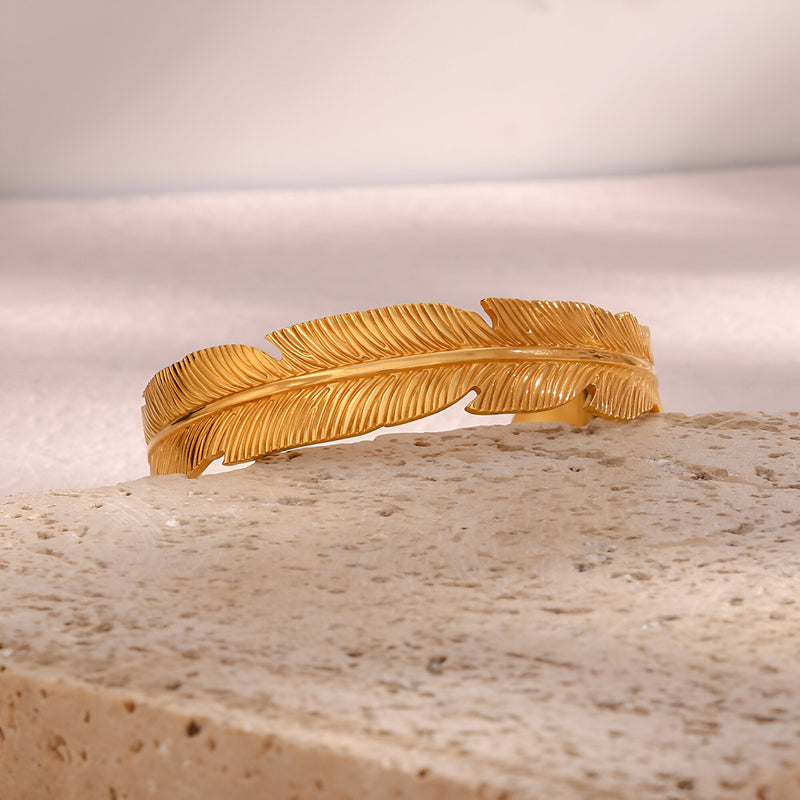 Bracelet jonc en acier inoxydable doré avec motif plume délicate et finition texturée.