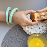 Bracelet jonc irrégulier en résine menthe porté au poignet féminin avec sandwich et verre de jus d'orange en arrière-plan.