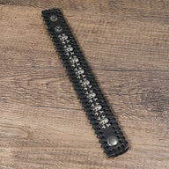 Bracelet large en cuir noir avec têtes de mort métalliques alignées et fermeture à bouton-pression.