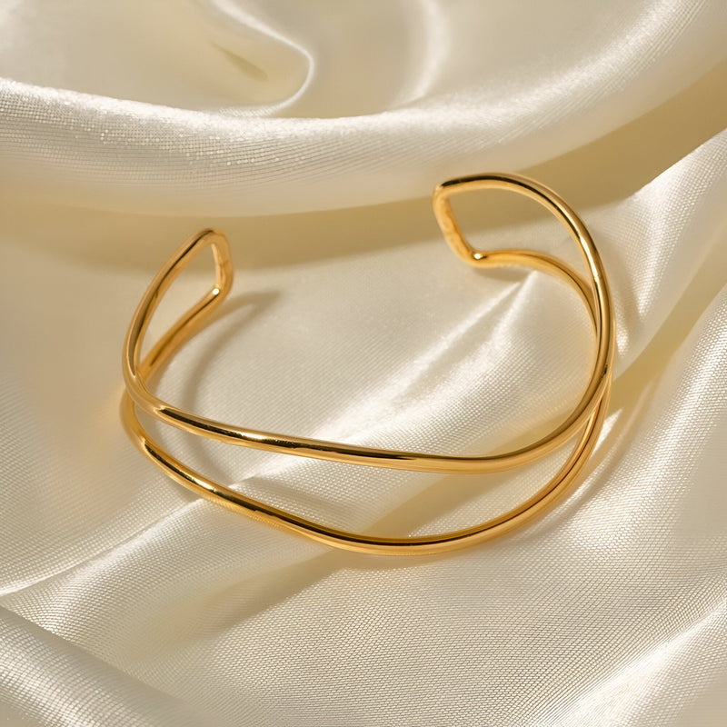 Bracelet manchette en titane plaqué or à design minimaliste et courbes élégantes sur fond satiné crème.