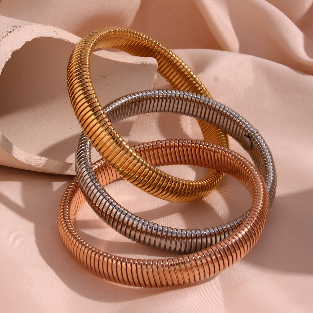 Ensemble trio de bracelets manchette en métal torsadé multicolore or, argent et cuivre brillant sur tissu rose pâle.