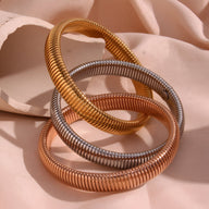 Ensemble trio de bracelets manchette en métal torsadé multicolore or, argent et cuivre brillant sur tissu rose pâle.