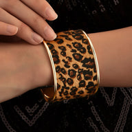 Bracelet manchette large à motif léopard brun et noir encadré de métal doré brillant sur poignet féminin.