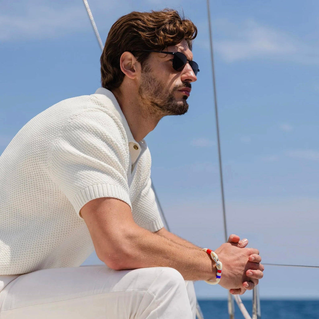 Bracelet mousqueton en coton beige avec drapeau français porté par un homme en pull blanc et lunettes noires sur un bateau.