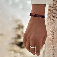 Bracelet multi-rangs en perles heishi violettes porté avec une bague argentée sur fond plage.