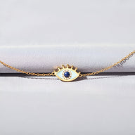 Bracelet femme en or avec pendentif œil serti d'une pierre bleue centrale et détails cloutés sur la paupière.