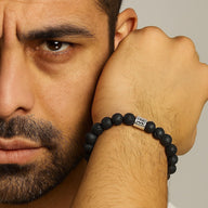 Bracelet élastique homme en perles rondes de pierre de lave noire avec breloque métallique argentée gravée de symboles.