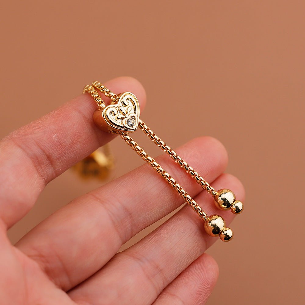 Bracelet ajustable doré à pendentif cœur gravé avec chaînes fines et boules aux extrémités.