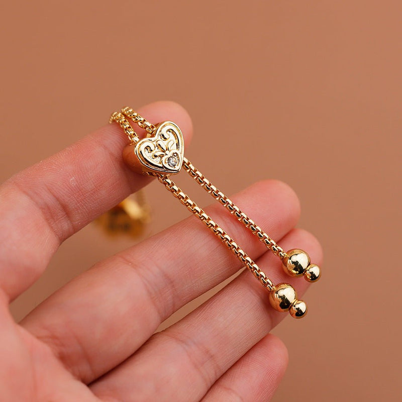 Bracelet ajustable doré à pendentif cœur gravé avec chaînes fines et boules aux extrémités.