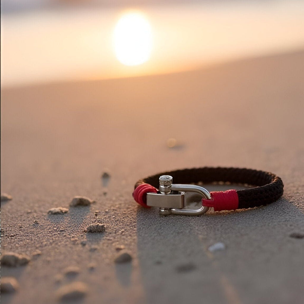 Bracelet homme en corde noire et rouge avec fermoir mousqueton argenté posé sur sable au coucher du soleil.