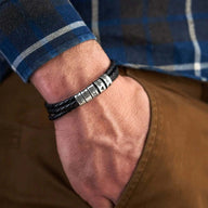 Bracelet en cuir noir tressé triple tour avec cinq anneaux métalliques gravables porté au poignet.