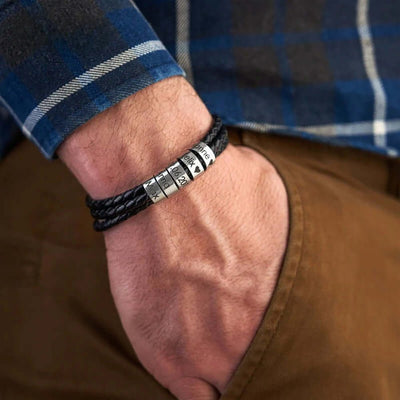 Bracelet en cuir noir tressé triple tour avec cinq anneaux métalliques gravables porté au poignet.