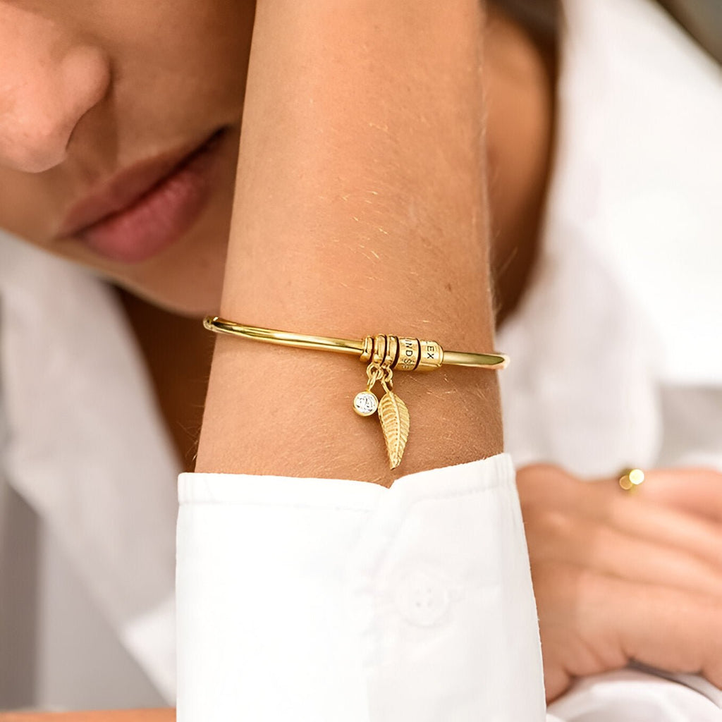 Bracelet fin deux anneaux en acier inoxydable doré avec pendentif plume et rond sur poignet féminin.