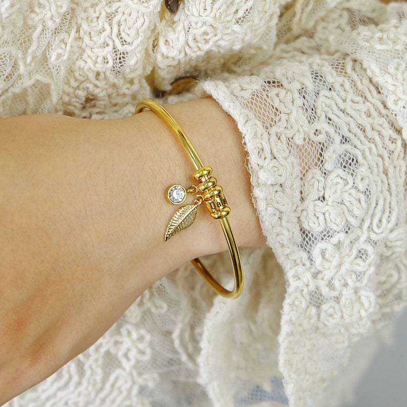 Bracelet fin en acier inoxydable doré avec anneau, feuille et pierre brillante, porté sur poignet féminin.