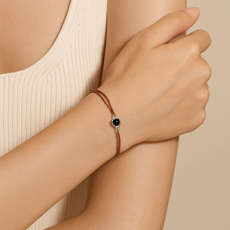 Bracelet fin à cordon cuir marron avec pendentif rond argenté et centre noir brillant.