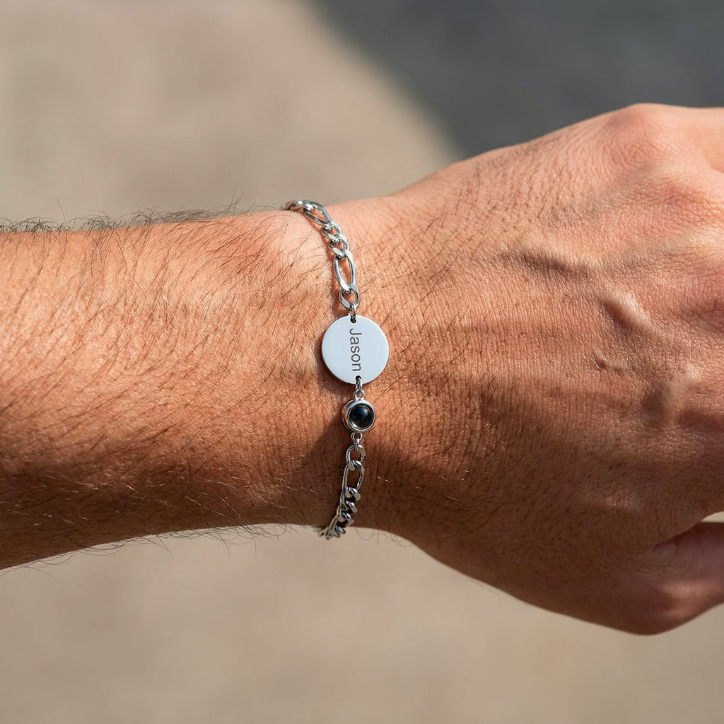 Bracelet en argent avec maille figaro, médaille ronde gravée et petit pendentif noir sur poignet masculin clair.