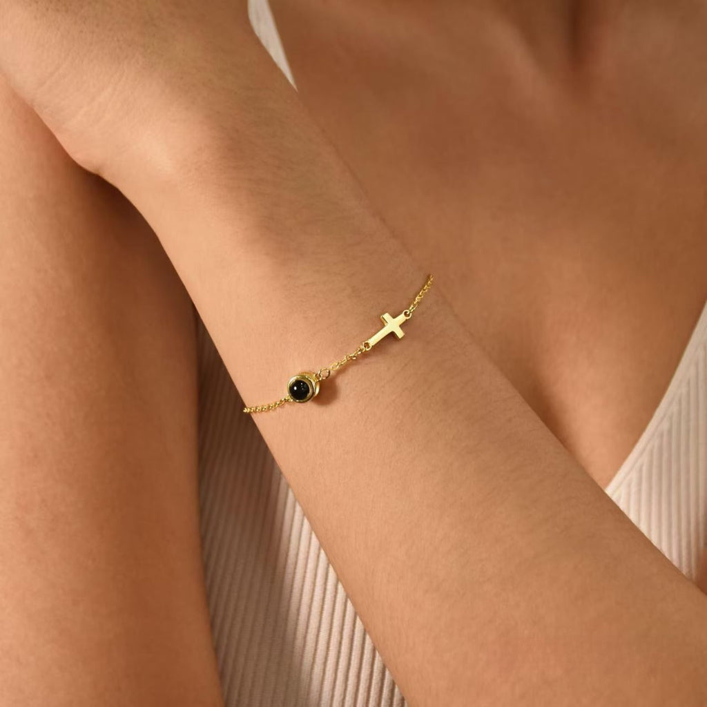 Bracelet fin en acier inoxydable doré avec une petite croix chrétienne et un médaillon circulaire noir porté au poignet.