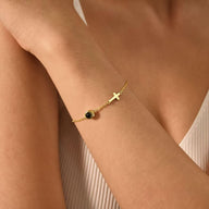 Bracelet fin en acier inoxydable doré avec une petite croix chrétienne et un médaillon circulaire noir porté au poignet.