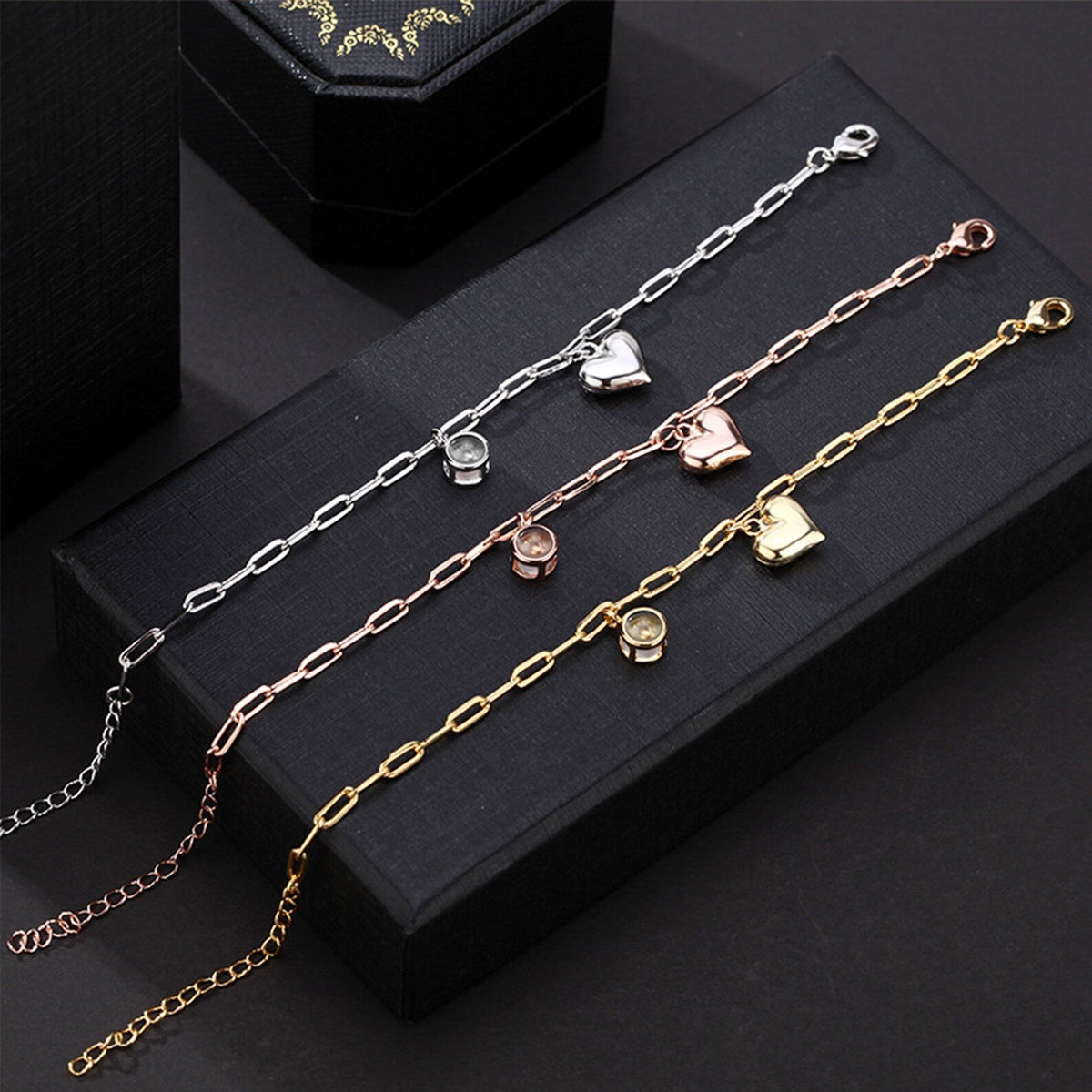 Trois bracelets personnalisés en titane avec charms cœur en or, argent et or rosé sur fond noir.