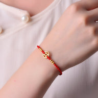 Bracelet en nylon rouge avec médaille dorée et perles dorées, style porte-bonheur, porté au poignet.