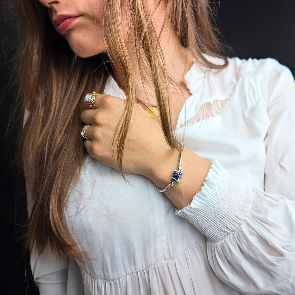 Bracelet argenté fin avec un charm papillon bleu délicat porté sur le poignet d'une femme en chemisier blanc.