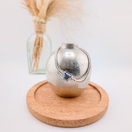 Bracelet en métal argenté avec pendentif papillon bleu serti de pierres sur présentoir argent et plateau en bois clair.