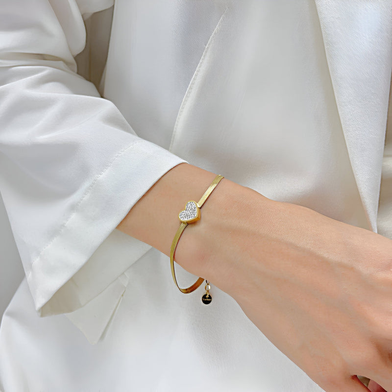 Bracelet fin doré en acier inoxydable avec cœur serti de zirconium porté au poignet féminin.