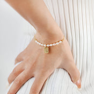 Bracelet doré pour femme en acier inoxydable avec perles blanches et pendentif rectangulaire gravé.