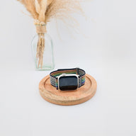 Bracelet Apple Watch en nylon noir avec rayures colorées et fermeture velcro, posé sur un plateau en bois rond.