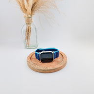 Bracelet Apple Watch en nylon bleu résistant avec fermeture velcro, modèle Elios, posé sur un support bois rond.