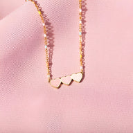 Bracelet en or avec trois cœurs alignés sur chaîne fine délicate posée sur tissu rose.