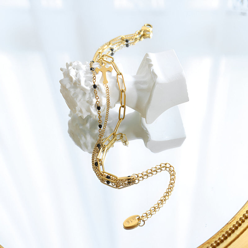 Bracelet triple chaînes en titane plaqué or avec motif croix et perles noires, fermoir ajustable.