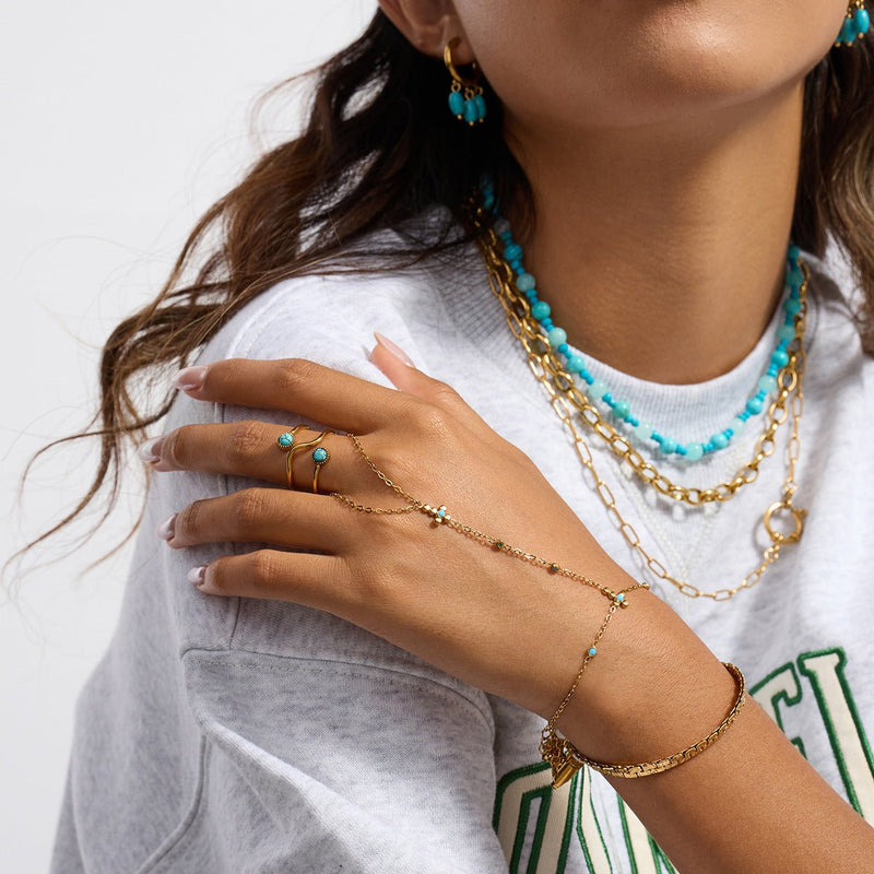 Chaîne de doigt en acier inoxydable doré avec perles turquoises et croix chrétienne délicate, portée sur main féminine.